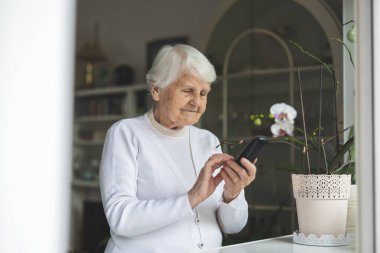 Evdeki oturma odasında cep telefonu kullanan yaşlı kadın.