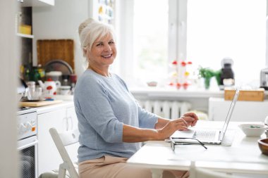 Dizüstü bilgisayarı ve defteri olan yaşlı bir kadın mutfaktaki masada oturuyor.