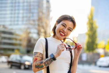 Kollarında dövmeleri olan güzel genç bir kadın şehirde gülümsüyor.