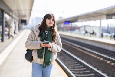 Cep telefonu olan genç bir kadın platformda tren bekliyor.
