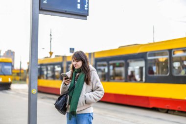 Şehirdeki tramvay durağında cep telefonu kullanan genç bir kadın.
