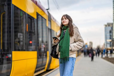 Şehirdeki tramvay durağında cep telefonu kullanan genç bir kadın.