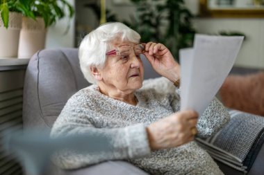 Oturma odasında bir koltukta otururken gözlük takan yaşlı kadın belgeleri okuyor.
