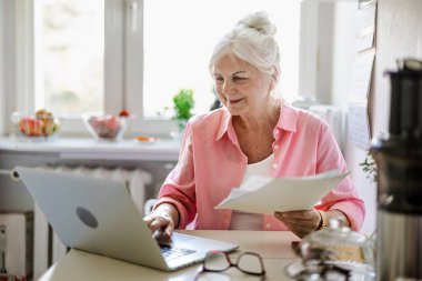 Üst düzey kadın evindeki mutfak masasında dizüstü bilgisayarla ve belgelerle çalışıyor.