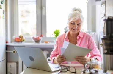 Üst düzey kadın evindeki mutfak masasında dizüstü bilgisayarla ve belgelerle çalışıyor.