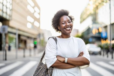 Güneşli bir günde şehirde duran gülümseyen genç bir kadının portresi.