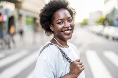 Güneşli bir günde şehirde duran gülümseyen genç bir kadının portresi.