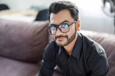 Sakallı adam evinin oturma odasındaki kanepede oturuyor ve kameraya bakıyor.