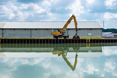 Yellow tractor crane loader on the bank of the canal with a reflection in the water.