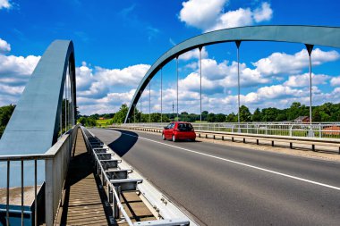 Arka planda bariyer ve köprü olan kırmızı araba yolda gidiyor..