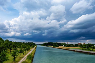 Nehir bulutlu gökyüzünün altında akar, bir tarafında ağaçlar, diğer tarafında köprüler vardır..