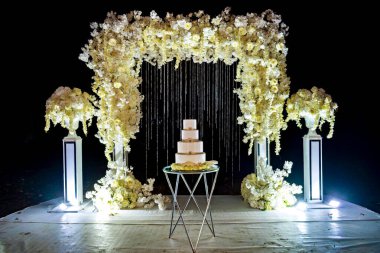 Düğün pastası masanın üstünde, çiçekli kemerin altında..