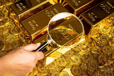 a hand with a magnifying glass checks the gold bar on a pile of gold coins clipart