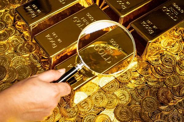 a hand with a magnifying glass checks the gold bar on a pile of gold coins
