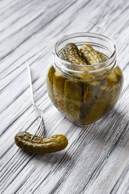 Pickled gherkins in an open glass jar and one cucumber on a fork nearby on a light wooden background close-up. Homemade pickled cucumbers.
