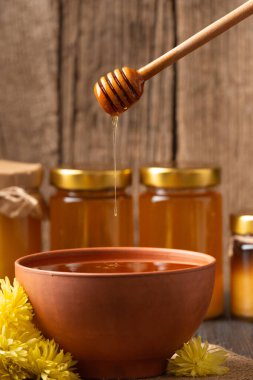 Beautiful drops of honey pour from a wooden dipper into a clay bowl with honey on a wooden background close-up. Composition of pouring honey, honey spoon, glass jars with honey and flowers.