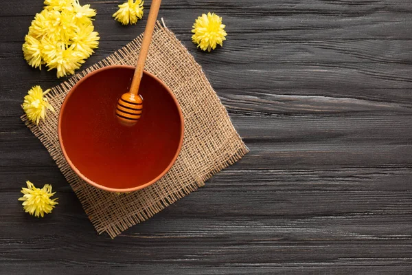 Honey in a clay bowl, dipper and yellow flowers on a textile background. A stick for honey lies in a clay bowl with honey top view with place for text . Composition of honey in a plate and flowers on a dark wooden background copy space.