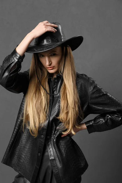 Pretty young sexy fashionable sensual girl posing beautifully on a dark background in a black leather shirt and a cowboy hat, clothing advertisement, a woman in a black leather hat.
