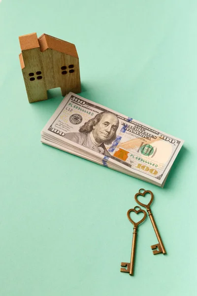 The concept of mortgage, purchase, rental housing, auction, loan, sale of real estate. Wooden house model, keys, stack of 100 dollars banknotes on a green background.