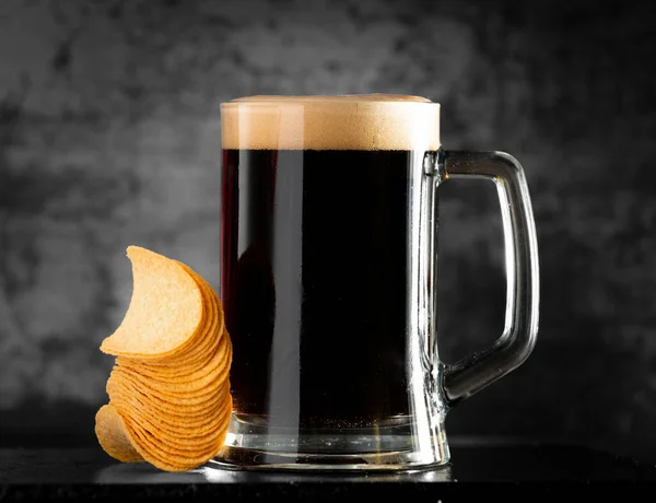 A glass of dark beer with a handful of chips on a dark background. Beer pouring from a bottle into a glass. Chips in a stack near a mug of beer with foam.