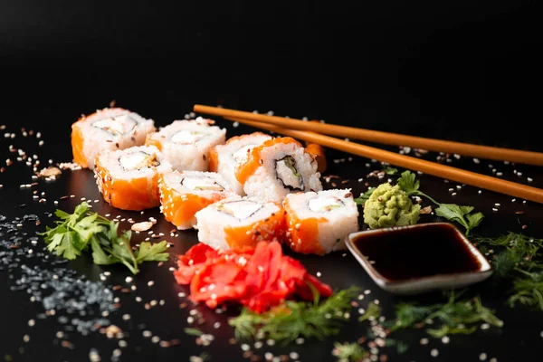 Set of salmon rolls on a dark background with chopsticks, sauce and red ginger. Red sushi with sesame on black background