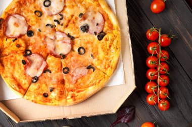 Açık karton bir kutuda pizza, soslar, baharatlı zeytinyağı, koyu ahşap arka plan manzaralı taze sebzeler. Yemek teslimatı.