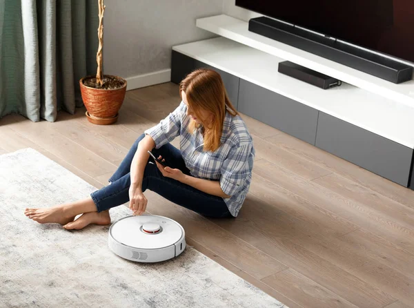 Bir kız oturma odasında yerde oturuyor ve akıllı telefon kullanarak robot elektrik süpürgesini kontrol ediyor. Akıllı Ev. Akıllı ev aletleri, ev işleri ve teknoloji kavramı..