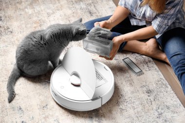 Elektrikli süpürge robotunun kabı kadın ellerindedir, robot elektrik süpürgesinin toz toplayıcısı yün doludur, kedi yakınlarda oturur ve konteynıra bakar..