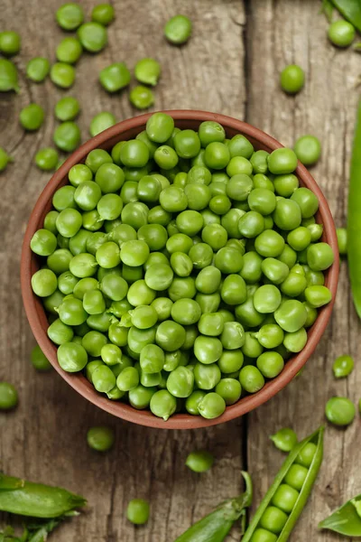 Fresh organic green peas in closed and open pods, scattered pea seeds, shelled green peas in a clay bowl on an aged wooden background, top view. vegetable protein.