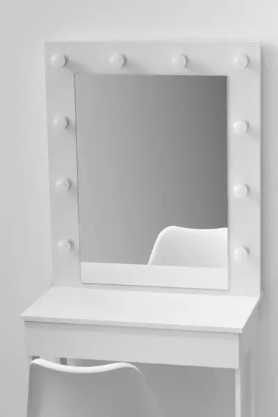 White dressing table for make-up with a large mirror and lamps, a white chair against the background of the wall, close-up. Makeup artist workplace, modern dressing room.