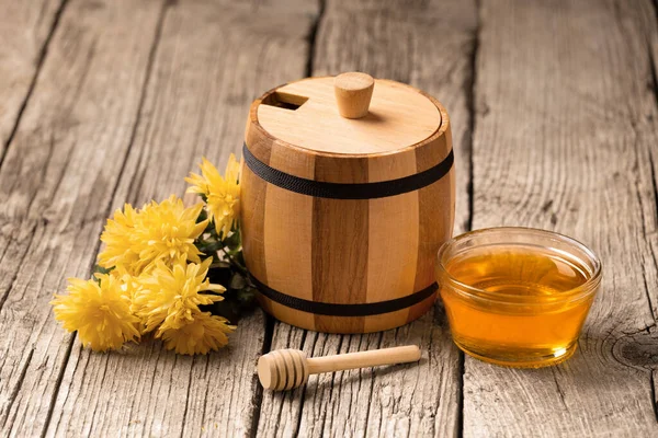 Honey in a small wooden barrel and a transparent glass bowl, a wooden dipper, yellow flowers on an aged wooden background, close-up.