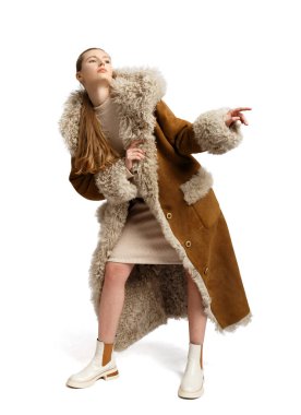 Kürk kapüşonlu ve kelepçeli uzun kahverengi kuzu derisi ceketli şık bir kız beyaz arka planda güzel poz veriyor. Modaya uygun, şık, rahat dış giyim..