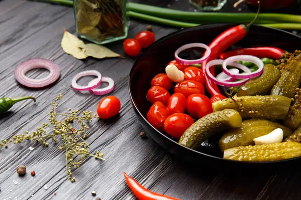 Assorted pickled vegetables. Canned tomatoes, cucumbers, chili peppers in a round black plate on a dark background. appetizer of canned vegetables. home preservation.