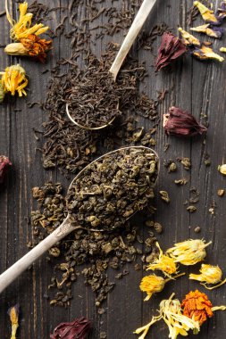 Antika kaşıklardaki kuru siyah ve yeşil çay kompozisyonu, koyu ahşap arka planda yakın plan görüntüsünde kuru çiçek çayı. Koyu arkaplanda farklı türde kuru çay..