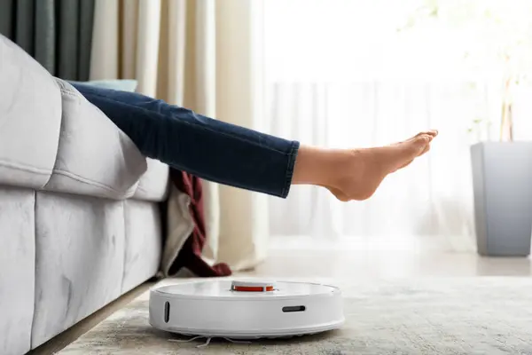 The robot vacuum cleaner does the cleaning in the living room. The girl raised her legs to let the robotic vacuum cleaner pass. The concept of smart home, technology, housework.