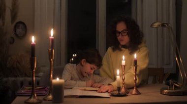 Kıvırcık saçlı anne mum ışığında elektriksiz karanlık bir dairede kızına ev ödevini yapmasına yardım ediyor.