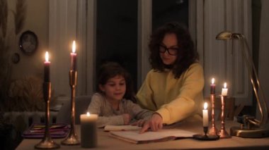 Kıvırcık saçlı süveter ve gözlüklü anne mum ışığında kızının ödevlerine yardım ediyor.