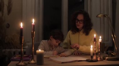 Kıvırcık saçlı süveter ve gözlüklü anne mum ışığında kızının ödevlerine yardım ediyor.