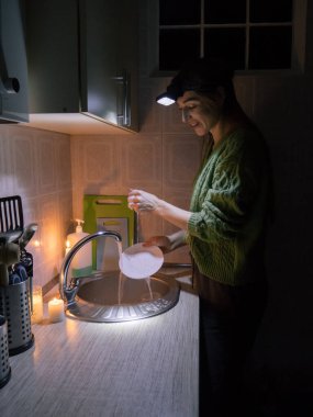 a young woman washes dishes in a kitchen without electricity in a sweater, and with a flashlight on her head by candlelight.
