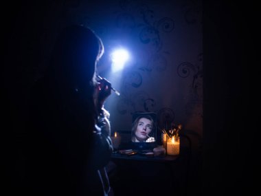 a woman holds a flashlight in her hands to illuminate herself when she does makeup in a room without electricity