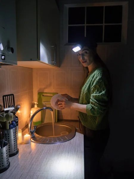 a young woman washes dishes and looks at the camera in a kitchen without electricity in a sweater, and with a flashlight on her head by candlelight.