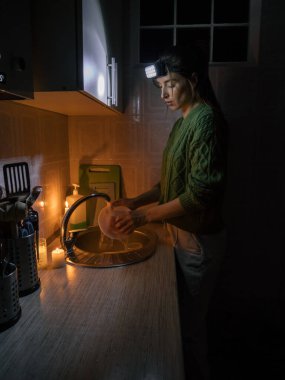 a young woman washes dishes in a kitchen without electricity in a sweater, and with a flashlight on her head by candlelight.