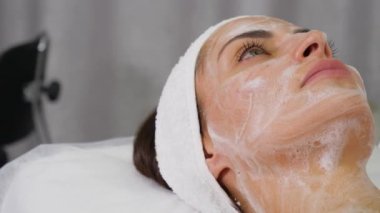 Yüzünde köpükle yatıp güzellik uzmanını bekleyen genç bir kadının yakın çekimi.