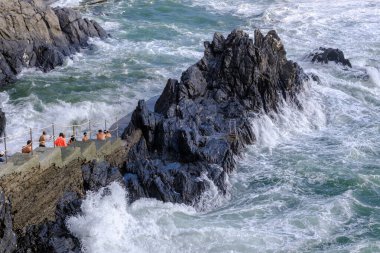 Manarola, IT - 25 Temmuz 2023: Gençler bir kayanın üzerinde kalırken büyük dalgalar kıyıya vuruyor