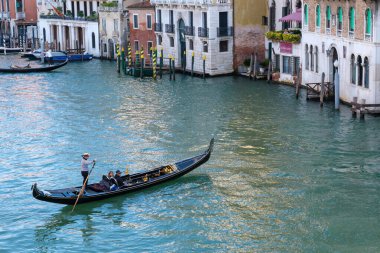 Venedik, İtalya - 12 Ekim 2024: Geleneksel bir Venedik gondolu, çizgili tişörtlü bir gondolcuyla Büyük Kanal 'da süzülerek iki yolcuyu tarihi binalar ve liman mimarisinden geçirdi