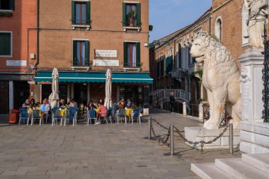 Venedik, İtalya - 12 Ekim 2024: İnsanlar Campo de l 'Arsenal' de açık bir kafede arka planda mermer bir aslan heykeli ile yemek yiyor