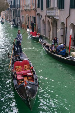 Venedik, İtalya - 12 Ekim 2024: Çizgili üniformalı bir gondolcu kırmızı koltuklu geleneksel bir gondolun başında duruyor