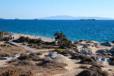 Naxos, Yunanistan - 3 Ekim 2024: Naxos 'taki Agios Anna Çıplaklar Plajı, birkaç güneş banyosu, şemsiye ve açık mavi gökyüzü altında tepelerin yer aldığı sakin bir kıyı şeridi