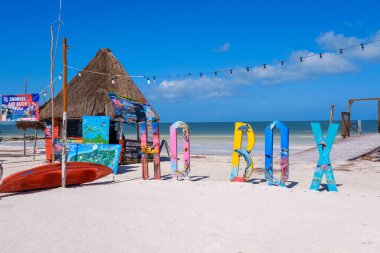Holbox, Meksika - 9 Şubat 2025: Turkuaz deniz manzaralı bir kano ve gezi posterleriyle birlikte plajda renkli Holbox tabelası.