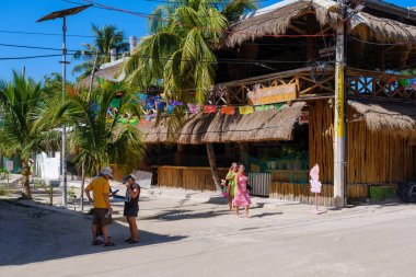 Holbox, Meksika - 9 Şubat 2025: Güneşli bir günde turistlerin sessiz bir yürüyüş yaptığı Holbox 'ta palmiye ağaçları ve sazlıklı çatı restoranları olan renkli cadde.